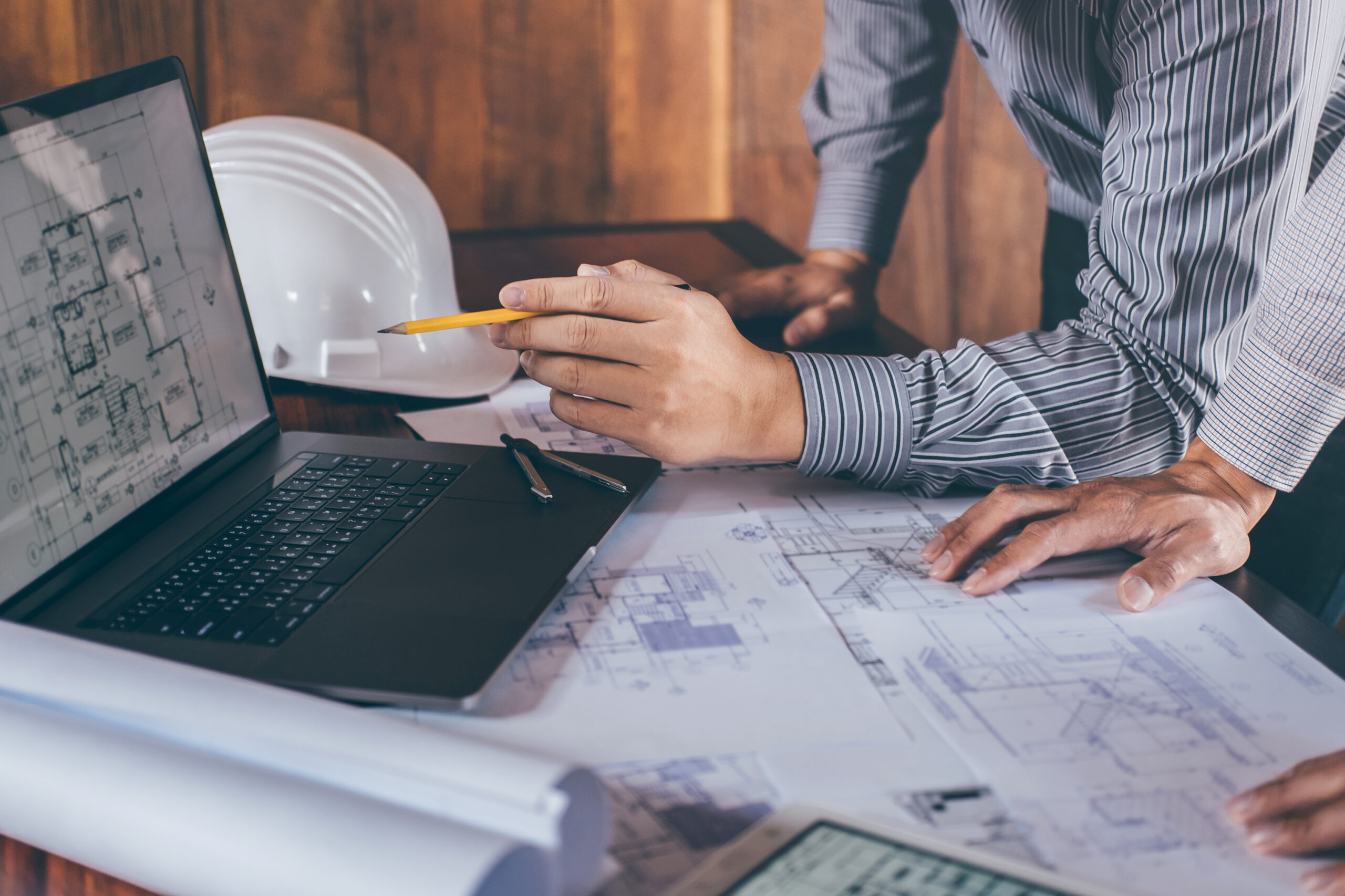 Odborníci konzultují pasportizaci budov a BIM modelování nad technickými výkresy a digitálním modelem z mračna bodů zobrazeným na notebooku. Na stole jsou rozložené plány stavby, v pozadí je viditelná ochranná přilba.